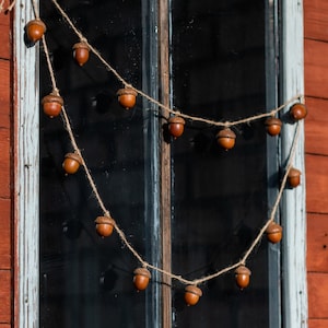 Puede incluir: Una guirnalda de bellotas marrones cuelga de un marco de ventana. La ventana es vieja y tiene un marco pintado de blanco. El fondo es una pared de madera roja.