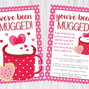 May include: Two pink and white Valentine's Day cards with a mug of hot chocolate and a heart-shaped cookie. The text on the cards reads "we've been mugged!" and "you've been mugged!" with instructions on how to play the game.