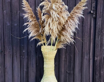 Op de afbeelding: Een boeket gedroogd pampasgras in een lichtbeige gewoven vaas. De vaas staat op een geweven tafelblad voor een donkerbruine houten muur.