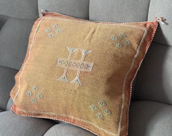 May include: A tan throw pillow with a geometric pattern in white and yellow thread. The pillow has a red border and is made of a soft, woven fabric.