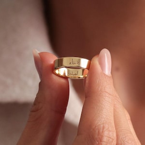 Puede incluir: Dos anillos dorados con texto grabado se sostienen frente a una persona. Los anillos tienen un diseño de banda plana y son de color dorado brillante y brillante. El fondo está borroso, con una camisa blanca y un collar dorado visibles.