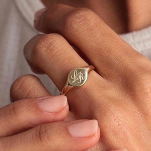 May include: A gold signet ring, featuring the engraved initials "LR" on its oval face. The ring is worn on a finger, highlighting its polished surface and traditional style. Another gold ring is visible on another finger.