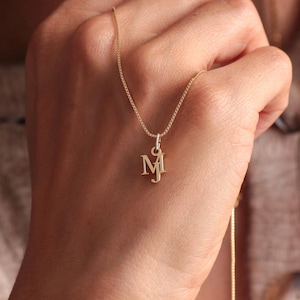 Op de afbeelding: Een goudkleurige ketting met een fijne ketting en een hanger met de letter "M". De hanger is in een gestileerd lettertype. De ketting wordt door een persoon vastgehouden.