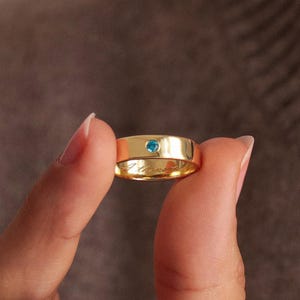 Peut inclure: Une bague anneau en or ornée d'une petite pierre précieuse ronde d'un bleu vif sertie au centre de la surface extérieure. Le métal présente une finition polie et l'anneau comporte une inscription gravée visible sur sa circonférence intérieure.
