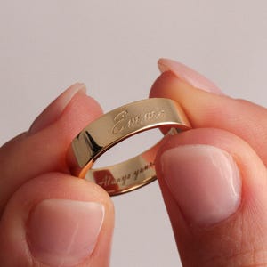 Puede incluir: Un anillo dorado con el nombre "Emma" grabado en el exterior. El anillo está sostenido entre dos manos. Las palabras "Always yours" están grabadas en el interior del anillo.