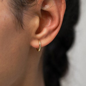 Puede incluir: Primer plano de un pendiente de aro dorado en una oreja. El pendiente tiene un diseño circular simple con un acabado pulido. La oreja es visible, mostrando la colocación y el tamaño del pendiente.