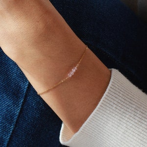 Könnte beinhalten: Ein zartes goldenes Armband mit kleinen, hellrosa Perlen. Das Armband wird an einem Handgelenk getragen, vor dem Hintergrund eines weißen Pullovers und einer blauen Jeans. Der Schmuck ist schlicht und elegant, passend für den Alltag.