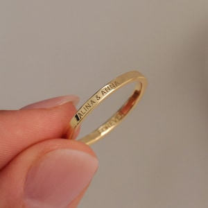 Puede incluir: Un anillo delgado de color dorado con las inscripciones grabadas "ALINA & ANNA" y "FOREVER". El anillo se sostiene entre los dedos sobre un fondo neutro, mostrando su delicado diseño e inscripción.