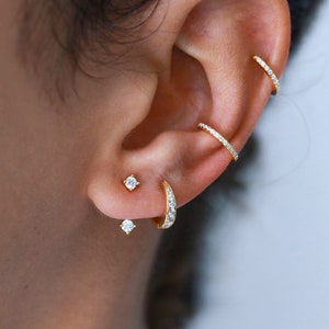 Op de afbeelding: Een oor met meerdere gouden oorringen. De oorringen zijn bezet met kleine, heldere stenen. De oorringen zitten in de helix-, conch- en lobe-piercings.