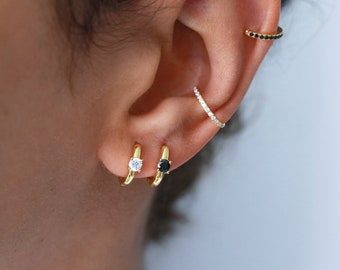 Puede incluir: Una oreja con varios aros de oro. Los aros están adornados con pequeñas piedras preciosas transparentes y negras.