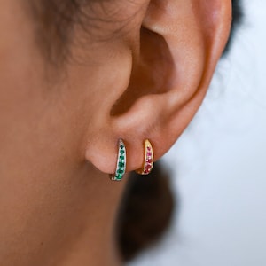 May include: Two small hoop earrings with a row of gemstones. The left earring has green gemstones and the right earring has red gemstones.