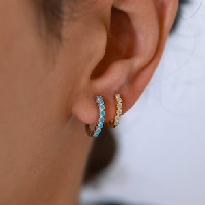 Peut inclure: Deux boucles d'oreilles créoles avec une rangée de pierres turquoise. La boucle d'oreille de gauche est en argent et celle de droite est en or.