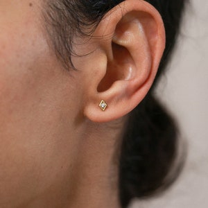 Può includere: Primo piano di un orecchio con un piccolo orecchino a perno di diamanti dorati. L'orecchino ha un'unica pietra trasparente al centro.