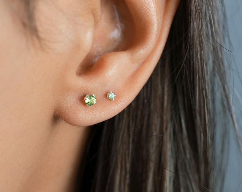 May include: A pair of small green gemstone earrings in silver settings. The earrings are worn in the lobes of a person's ear.