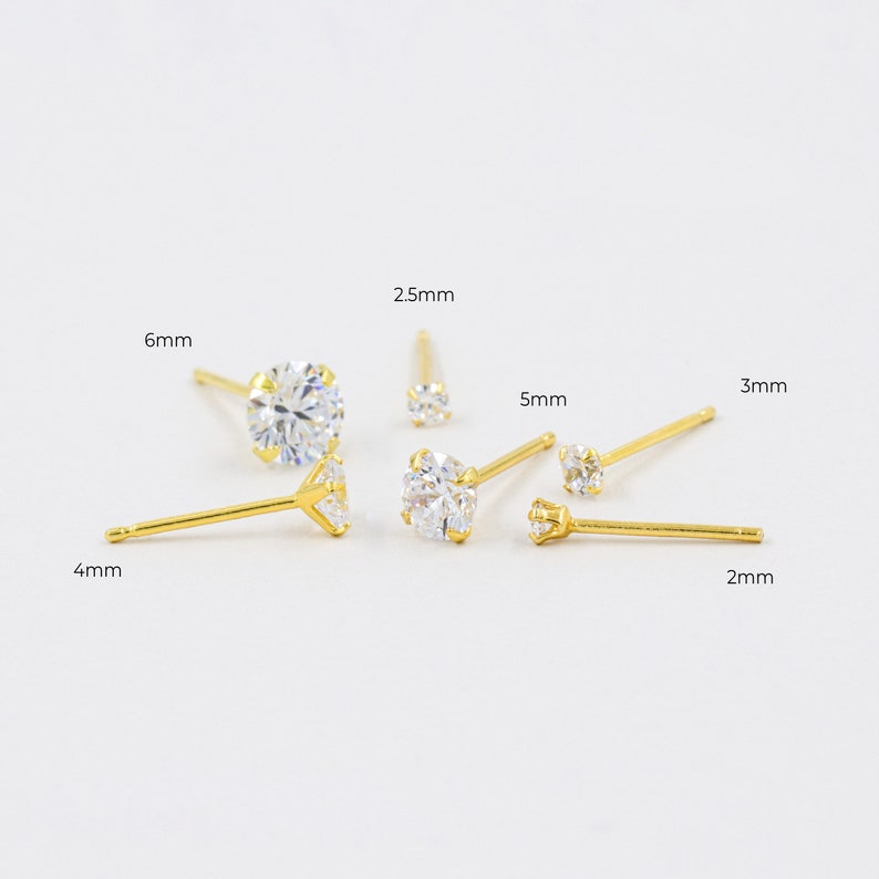 May include: A collection of gold-colored stud earrings with clear, round cubic zirconia stones. The earrings vary in size, with measurements of 2mm, 2.5mm, 3mm, 4mm, 5mm, and 6mm. The earrings are displayed on a white background.