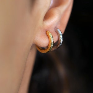 Puede incluir: Un par de pendientes de aro martillados, uno dorado y otro plateado, puestos en la oreja.