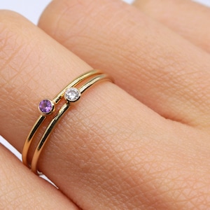 May include: Two gold rings with a small purple gemstone and a small clear gemstone on a finger.