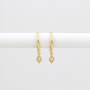 May include: A pair of gold hoop earrings with a dangling diamond-shaped charm. Each charm features a small, clear cubic zirconia stone.