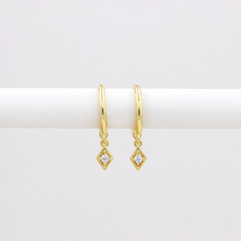 May include: A pair of gold hoop earrings with a dangling diamond-shaped charm. Each charm features a small, clear cubic zirconia stone.