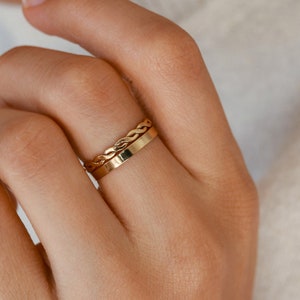 Peut inclure: Deux bagues dorées superposées sur un doigt. La bague du dessus a un motif tressé, tandis que la bague du dessous est une simple bande. Les bagues sont en or brillant.