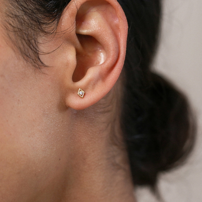 May include: A close-up of a person's ear with a small diamond stud earring. The earring is set in a gold frame and has a square shape.