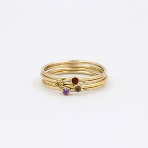 May include: Four gold stacking rings with small gemstones. The rings feature a yellow, red, purple, and gray gemstone.
