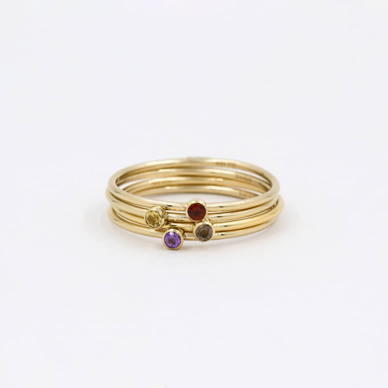 May include: Four gold stacking rings with small gemstones. The rings feature a yellow, red, purple, and gray gemstone.