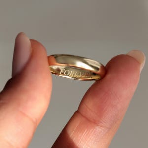 Puede incluir: Una alianza de boda dorada con la palabra "FOREVER" grabada en el interior. El anillo se sostiene entre dos dedos, mostrando su superficie pulida y su diseño sencillo. La banda tiene una forma lisa y redondeada.