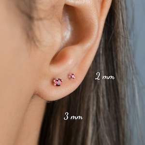 May include: Two pairs of small stud earrings with red gemstones. The earrings are shown in a person's earlobe. The left earring is 3 mm and the right earring is 2 mm.