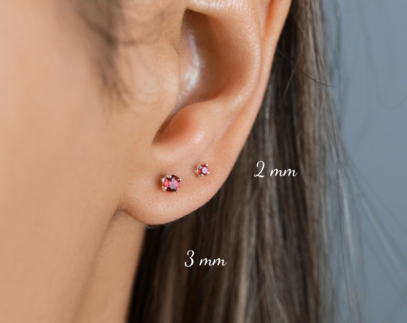 May include: Two pairs of small stud earrings with red gemstones. The earrings are shown in a person's earlobe. The left earring is 3 mm and the right earring is 2 mm.