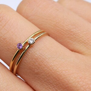 May include: Two gold rings with small gemstones. The top ring has a clear gemstone and the bottom ring has a purple gemstone.