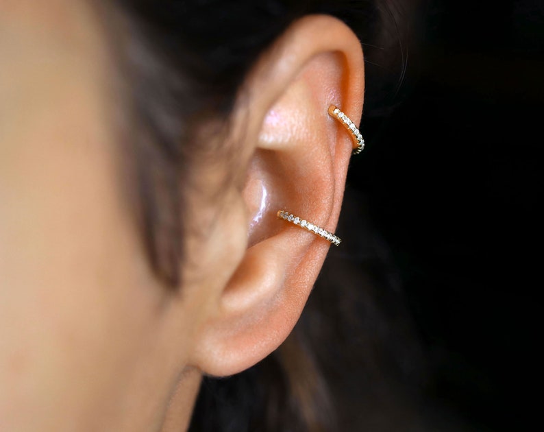 May include: Two gold ear cuffs with a row of small clear cubic zirconia stones. The cuffs are worn on the upper part of the ear.