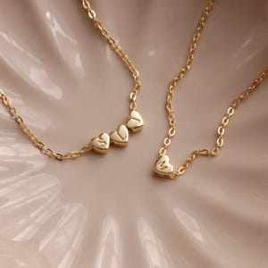 Peut inclure: Colliers dorés avec pendentifs en forme de cœur. L'un des colliers comporte trois cœurs avec les lettres "S", "V" et "L". L'autre collier a un seul pendentif en forme de cœur avec la lettre "M". Les colliers sont sur une surface rose clair.