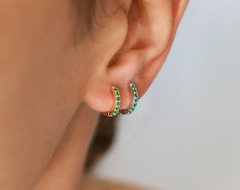 May include: A pair of small hoop earrings with a gold and silver finish. The hoops are set with small green gemstones.