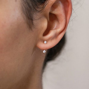 Peut inclure: Une paire de petites boucles d'oreilles délicates avec une seule pierre précieuse transparente sur chacune. Les boucles d'oreilles sont en or et ont un design simple et élégant.
