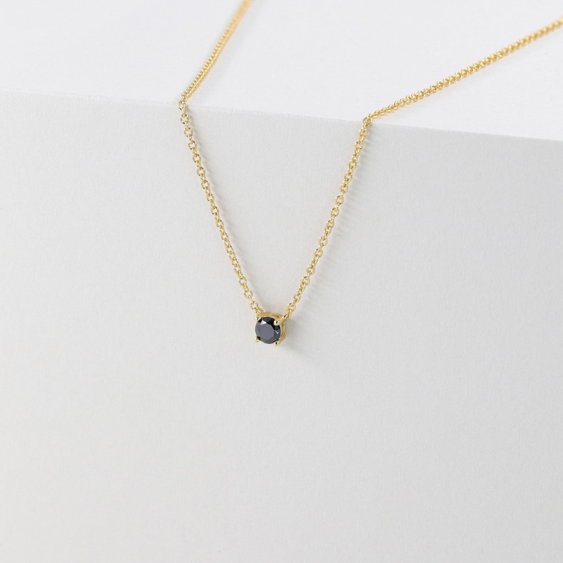 May include: A delicate gold necklace featuring a small, round, black gemstone pendant. The chain is fine and the pendant is set in a simple, minimalist style. The necklace is displayed against a white background.