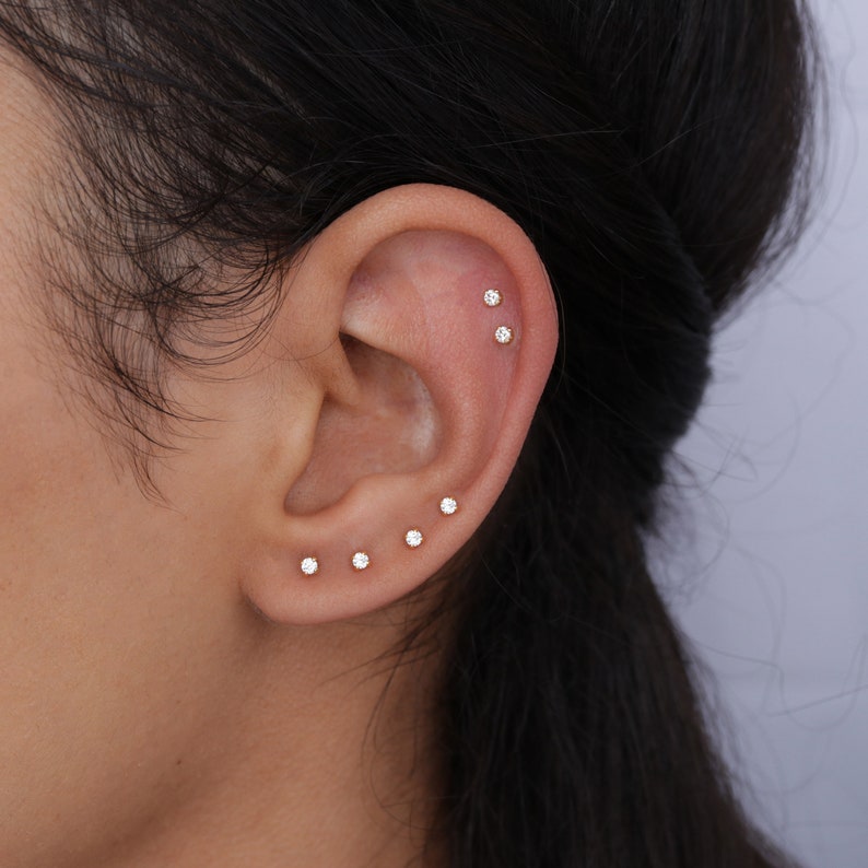 May include: Close-up of an ear with multiple small, gold-colored stud earrings. The earrings are evenly spaced along the ear's cartilage and lobe, each featuring a small, clear gemstone. The ear is set against a blurred background.