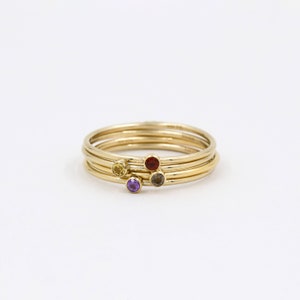 May include: Four gold stacking rings with small gemstones. The rings feature a yellow, red, purple, and gray gemstone.