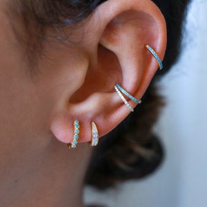 May include: A person's ear with multiple gold hoop earrings. The earrings are set with small turquoise and white stones.