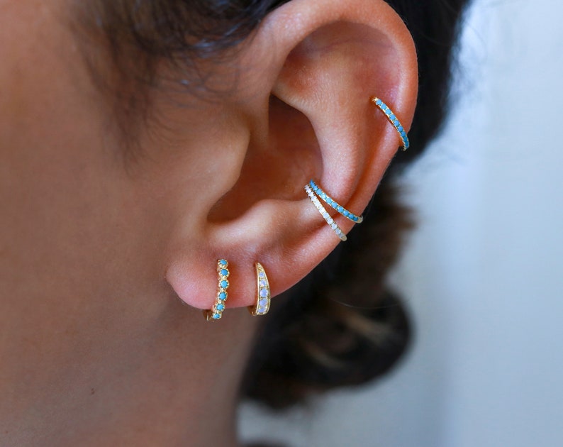 May include: A person's ear with multiple gold hoop earrings. The earrings are set with small turquoise and white stones.