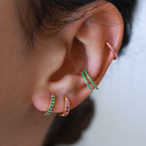 May include: Three gold hoop earrings with small green and red gemstones. The earrings are worn in the earlobe and helix piercings.