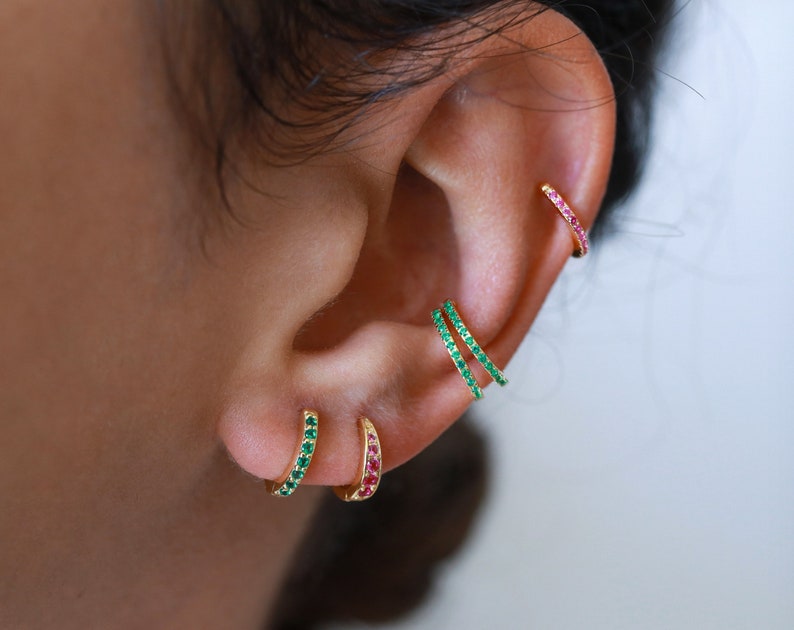 May include: Three gold hoop earrings with small green and red gemstones. The earrings are worn in the earlobe and helix piercings.
