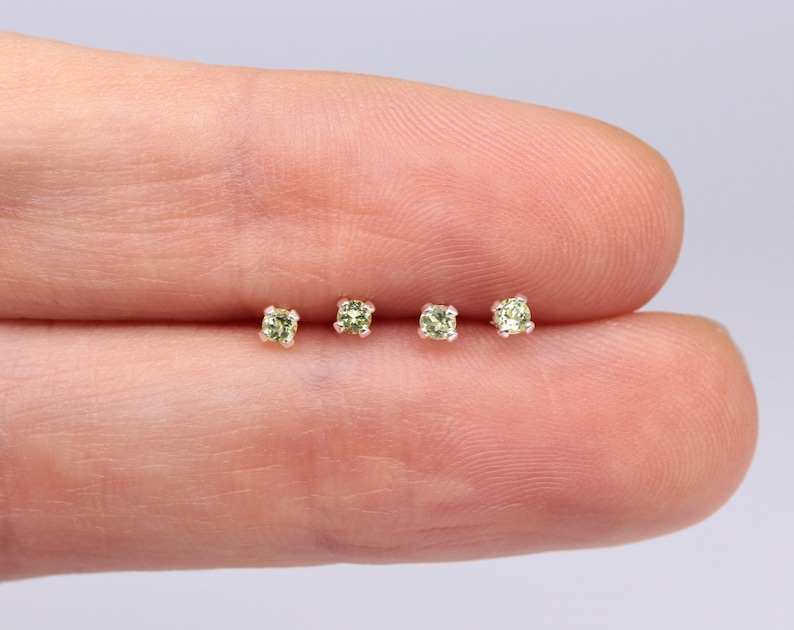 May include: Four tiny green gemstone earrings on a finger. The earrings are set in silver.