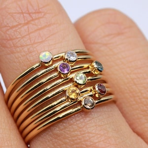 May include: A set of nine gold stacking rings with different colored gemstones. The gemstones include opal, white, amethyst, green, yellow, and red.