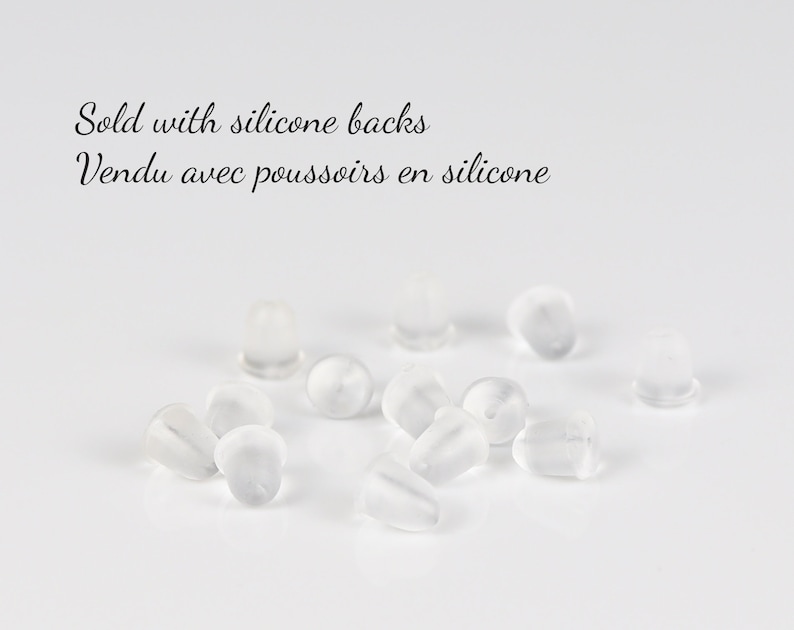 May include: A close-up of a pile of clear silicone earring backs. The text "Sold with silicone backs Vendu avec poussoirs en silicone" is visible in the image.