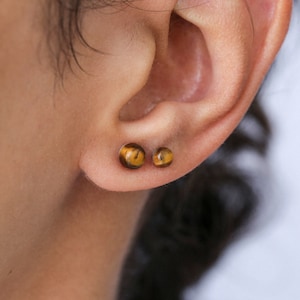 May include: Close-up of a person's ear with two small, round, brown and gold tiger's eye earrings.