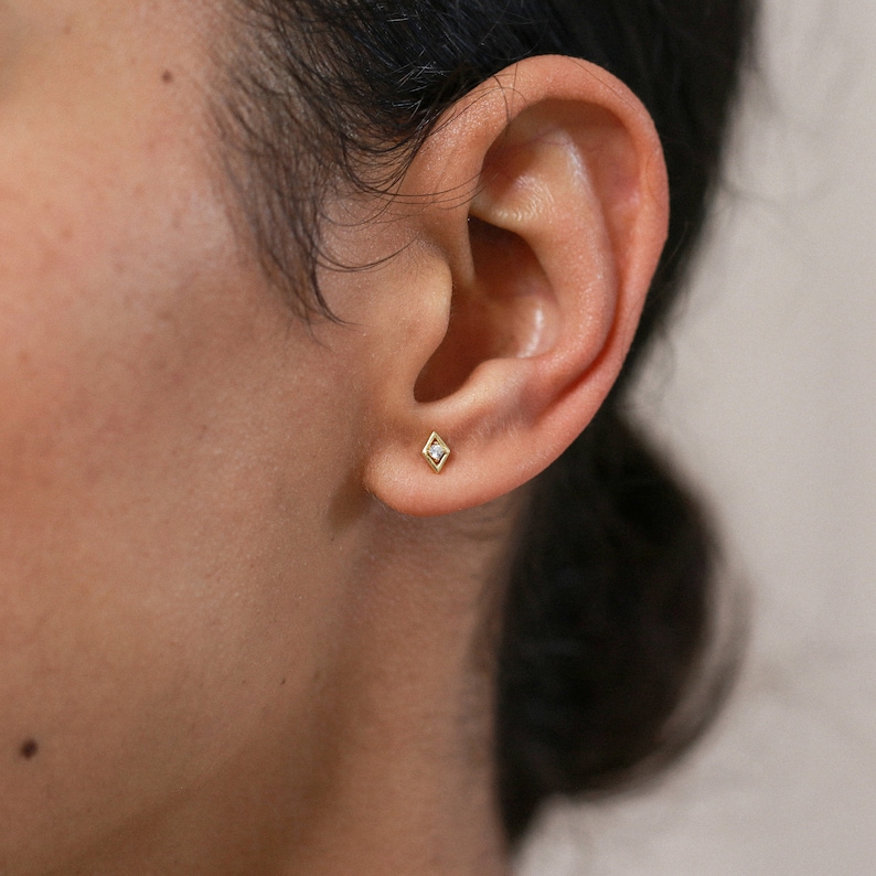 May include: A close-up of a person's ear with a small gold diamond earring. The earring has a single clear stone in the centre.