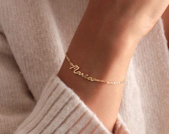 May include: A delicate gold-colored bracelet with the name "Maria" in cursive script. The bracelet is attached to a fine chain and is worn on a wrist, suggesting a personalized jewelry item.
