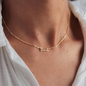 Puede incluir: Un delicado collar dorado con el nombre "Aria" en escritura cursiva. El collar se lleva alrededor del cuello y la cadena es visible. El fondo es una camisa blanca.
