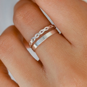 Peut inclure: Deux bagues en argent sur un doigt. La bague du haut est un modèle tressé et la bague du bas est une bande simple.